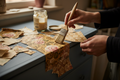 Hands applying torn vintage wallpaper pieces onto a painted wooden furniture surface using decoupage, warm moody workshop lighting